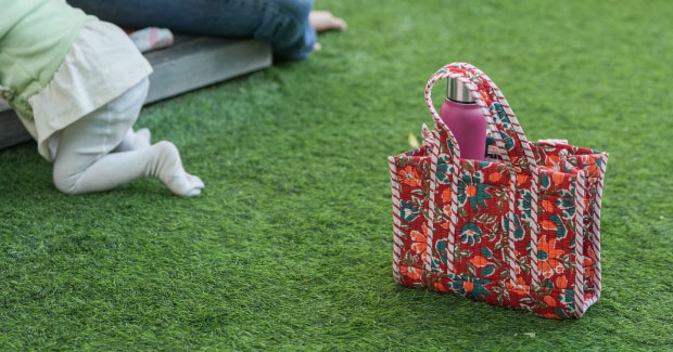 Red floral printed Jaipur cotton tote bag holding a water bottle on grass, a reusable cloth bag by Navo Juno Lifestyle